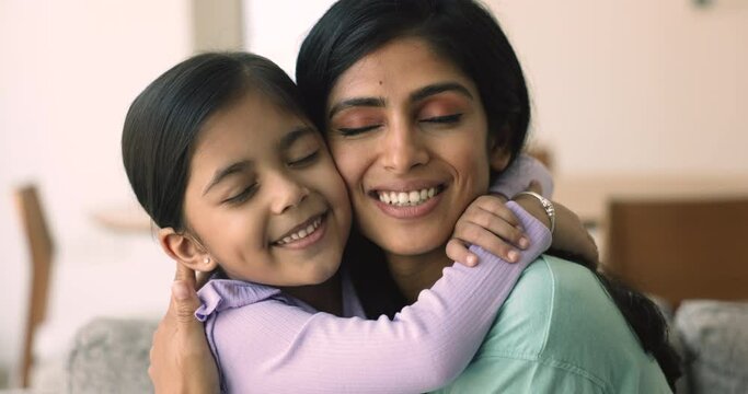 Close up beautiful peaceful faces of little 5s Indian girl cuddling tightly her young loving mommy with eyes closed, feeling unconditional love, enjoy moment of tenderness. Happy motherhood, adoption