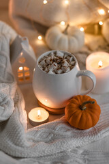 White mug with aromatic autumn hot drink - sweet delicious pumpkin latte with marshmallow and spices White background, bokeh lights, candles, knitted sweater as decor