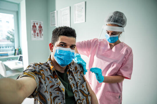 Portrait of a young man taking a selfie on a smartphone while getting vaccinated by a health worker at the hospital