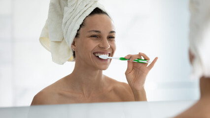 Smiling young woman in towel after shower look in bath mirror brush white healthy teeth with...