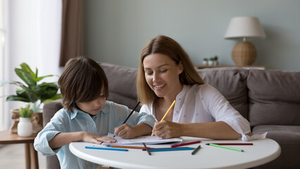 Happy beautiful mom teaching adorable little son to draw in coloring pencils, scratching doodles in paper albums at small table in living room, practicing artistic creative skill, educational activity
