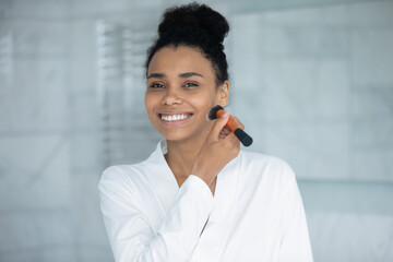 Portrait of beautiful smiling african American young woman get ready do make up in bathroom at home, happy biracial female use brush put powder beauty product on cheeks and face, cosmetics concept - Powered by Adobe