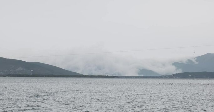 Lake Sevan, one of the largest freshwater high-altitude lakes, is captured on a day where the overcast sky allows clouds to graze the water's surface. The serene environment is accentuated by distant