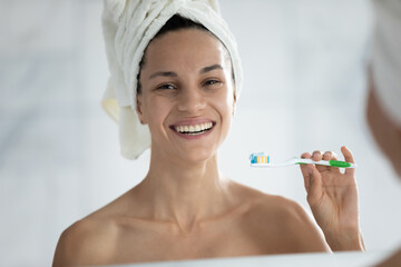 Portrait of smiling young Caucasian girl look in mirror in bathroom brush teeth with toothbrush and toothpaste, happy woman after shower clean do daily morning oral hygiene routine in home bath