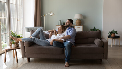 Happy peaceful married couple relaxing at home, resting on comfortable soft couch, enjoying leisure, dating, love relationship, hugging, looking away, thinking, dreaming