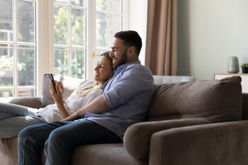 Positive millennial married couple relaxing on comfortable couch, using online app on mobile phone,...