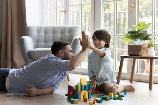 Happy father and little son boy playing constructing game on heating floor, celebrating toy model completion, giving high five, clapping hands over stacked cubes, blocks, enjoying family home activity