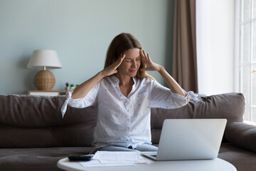 Exhausted stressed millennial laptop user woman touching head with closed eyes, feeling headache, sitting in sofa at paper documents, pc, calculator at home, feeling frustrated about too high fees