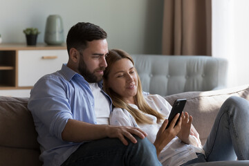 Calm relaxed millennial couple in love holding mobile phone on home sofa, sitting close, using...