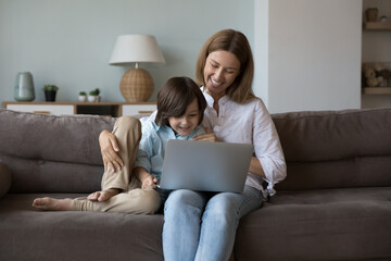 Cheerful beautiful mom and curious kid using online learning app on laptop for preschooler, watching cartoon movie, making video call, holding gadget for wireless connection, communication, laughing