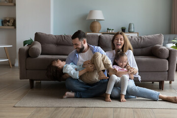 Excited joyful parents tickling energetic little kids on warm floor, laughing, having fun, enjoying family activity at home. Happy mom and dad playing with giggling, shouting son and daughter kids