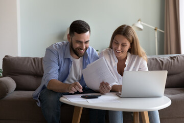 Happy satisfied millennial couple of renters, homeowners, bank clients counting rental, mortgage fees, calculating money, income, expenses, checking paper bills, doing domestic paperwork together