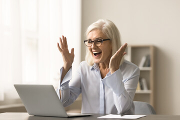 Cheerful blonde elderly business woman celebrating professional success, working at laptop, laughing, shouting for joy at home office workplace, getting good news from video call