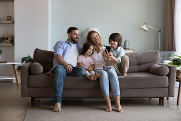 Happy millennial parents and two little children using mobile phone on home sofa, hugging, laughing, having fun, enjoying leisure, online communication, shopping on Internet