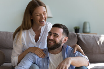 Fototapeta premium Cheerful attractive couple in love having fun at home, sitting close in loving room, talking, chatting, smiling, laughing, hugging with love, tenderness, enjoying leisure, relationship