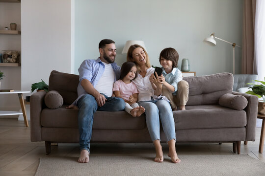 Happy millennial parents and two little children using mobile phone on home sofa, hugging, laughing, having fun, enjoying leisure, online communication, shopping on Internet