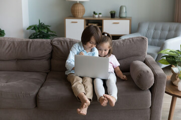 Focused curious sibling gen Z kids using laptop, holding computer, sitting on home couch, browsing Internet, blogs, chatting online, playing virtual games, enjoying wireless communication alone