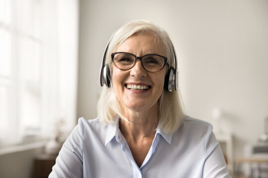 Happy Pretty Blonde Older Business Woman In Wireless Headphones And Glasses Speaking On Online Conference, Looking At Camera With Toothy Smile, Laughing. Video Call Screen Head Shot Portrait