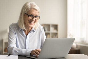 Cheerful blonde business woman in elegant glasses typing on laptop computer at home, sitting at workplace desk, enjoying online job communication, using wireless Internet technology, laughing - obrazy, fototapety, plakaty
