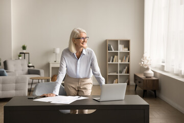 Happy successful senior freelance business woman standing at home workplace table with laptop,...