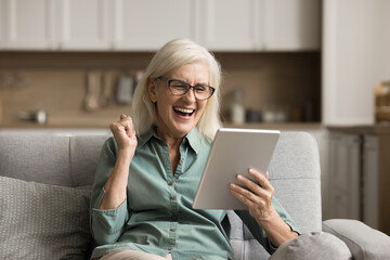 Cheerful excited elderly woman using digital tablet computer, making winner yes hand, laughing,...