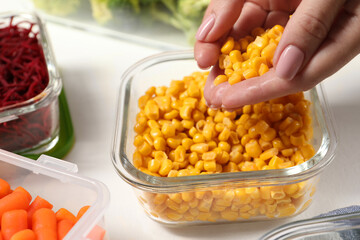 Woman putting corn into glass container at white table, closeup. Food storage
