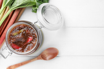 Jar of tasty rhubarb jam, stems and spoon on white wooden table, flat lay. Space for text