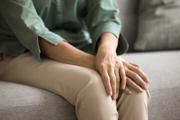 Mature elderly woman in casual sitting on couch, folding arms, wrists over knees, leaning elbows on hips, keeping closed posture, body gesture of waiting, concern. Female hands close up