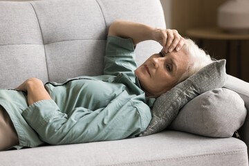 Depressed elderly woman resting on home couch, lying on back, looking away, touching head,...