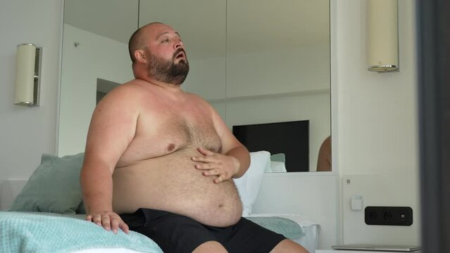 Fat guy suffers from summer heat. Chubby man breathes heavily, sweating profusely, wipes face and body with hand, regulates power of air conditioner, suffocates from abnormal heat and stuffiness