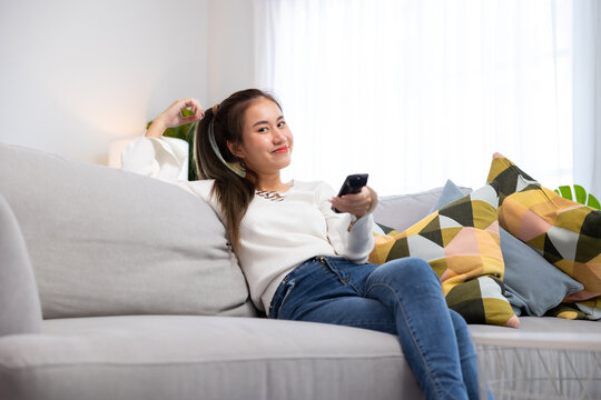 Leisure Watching TV and using remote controller in living at home. Relax, Asian thai young woman with remote and watching tv on sofa in living room. Entertainment