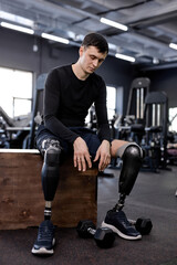 Fototapeta na wymiar sportsman is tired , sitting on the box, looking at dumbbells on the floor. full length photo .tiredness, modern sport equipment in the background of the photo