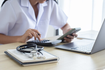 Female doctor or practitioner using calculator and mobile phone, work on laptop computer with medical stethoscope and notebook on the desk at clinic or hospital. 