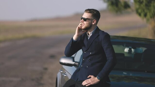 Successful businessman talking on phone sitting on car in park outdoor field road, smiling and looking to sun in stylish glasses