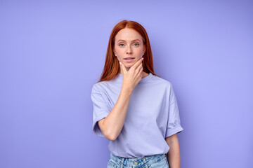 beautiful ginger girl with hand on her chin looking at camera, red-haired girl has idea, plan, touching her chin, close up portrait isolated blue background