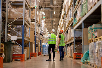 two men in warehouse wearing safety helmet, vest. Concept for industry, job, meeting, work training. Two caucasian warehouse workers walking in distribution storage area discussing - obrazy, fototapety, plakaty