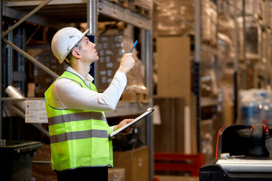 concentrated caucasian man worker in hard hat and working wear outfit doing stocktaking of products on shelves in warehouse using clipboard, taking notes. Physical inventory count concept
