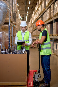 two warehouse managers walking through large warehouse distribution center discussing about increasing productivity. good teamwork, men dressed in working clothes vest and hard hats