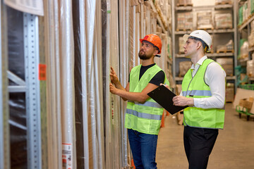confidentmale warehouse workers in working clothes talking together in warehouse, caucasian men counting goods in shelves, controlling the disctribution, industrial business concept