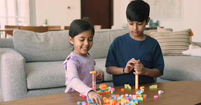 Little Indian boy and girl play plastic colorful built blocks together at home. Pretty 5s sister enjoy playtime with older 8s brother, smile talk having good, friendly relations. Play-time of siblings