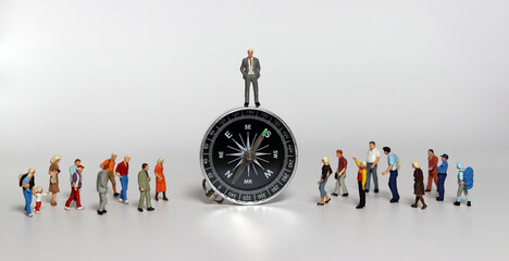 A miniature man is standing on a compass and miniature people are standing around him. Concepts...