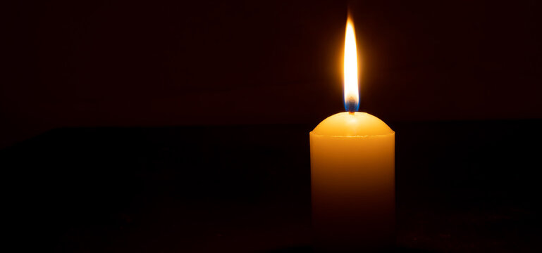 Single burning candle flame or light glowing on a big white candle on black or dark background on table in church for Christmas, funeral or memorial service with copy space.