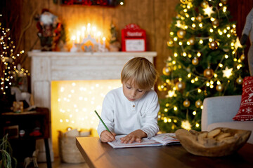 Fototapeta na wymiar Beautiful blond child, young school boy, writing homework for school in a decorated home, drinking milk and eating cookies. Ligths and knitted toys