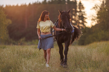 A young girl walks across a field and leads a horse. Rider against the backdrop of the forest at...