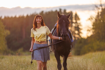 A young girl walks across a field and leads a horse. Rider against the backdrop of the forest at...