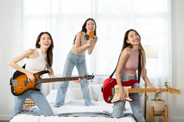 Beautiful three young Asian girls having fun singing and dancing in bedroom. Happy group of Asian friends playing musical instruments guitar bass at home. Friendship leisure rest home enjoy concept