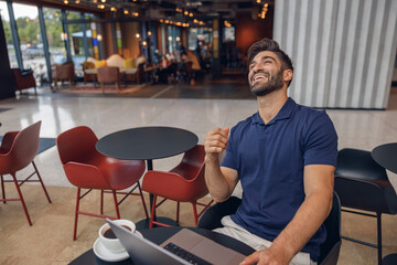 Happy male freelancer laughs during work on laptop in cozy cafe. Distance work concept