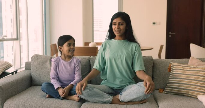 Little playful girl and her young mom meditating together seated on sofa in living room at home. Loving mother teach yoga practice daughter. Lifestyle, good life habit of Indian family, mindfulness