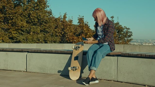 Trendy cool pretty pink haired female skateboarder sitting on bench, taking photo of skateboard wheel on cellphone and browsing online for spare part while skateboarding in urban settings at dawn.