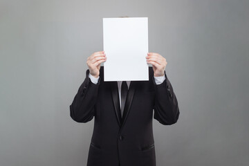 Business person wearing black suit hiding his face behind blank empty white paper on gray studio wall background
